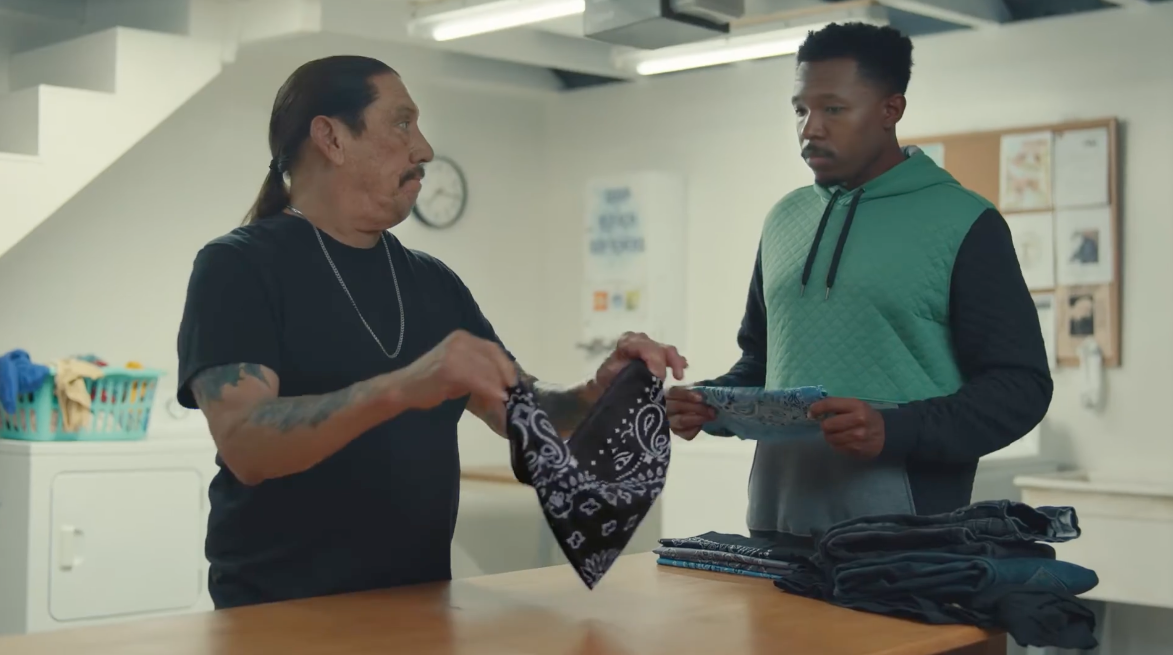 Danny Trejo holds a black bandana while looking over at a man holding his laundry